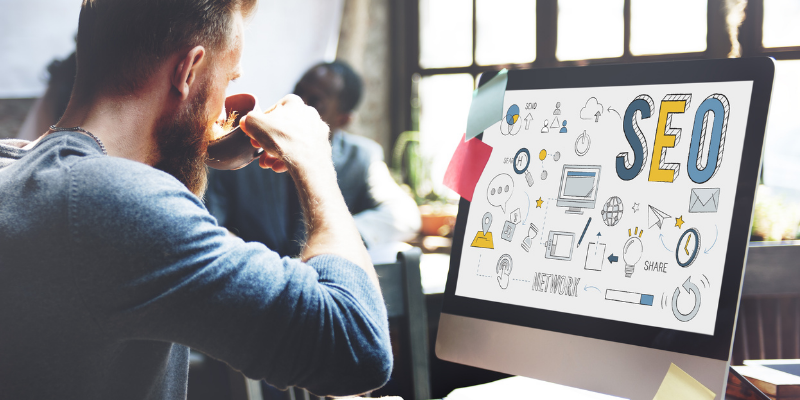 Hombre tomando de su taza de cafe frente a una computadora con un fondo de pantalla que dice SEO.